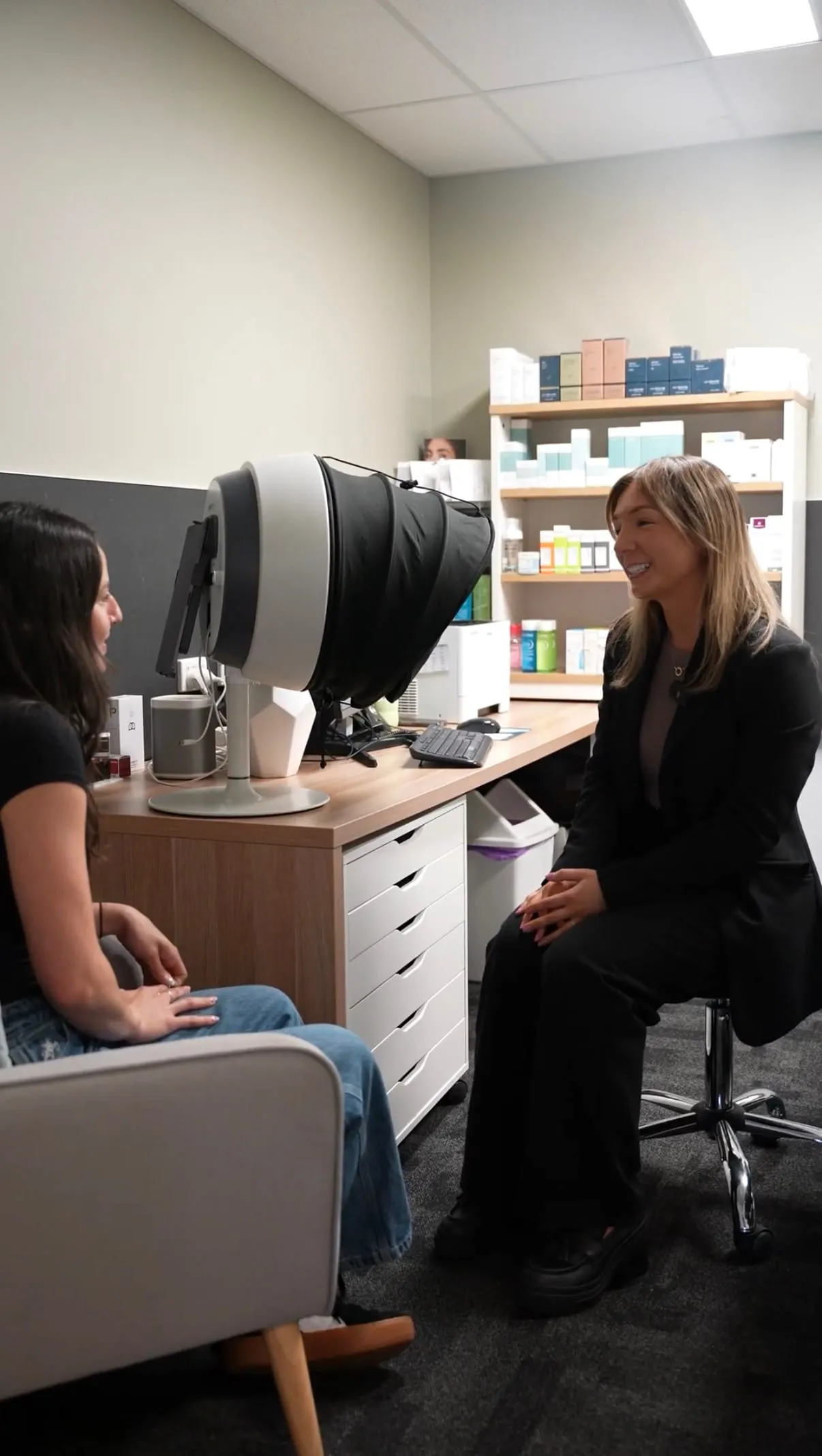 A dermal clinician consulting a patient beside the Observ skin-analysis system, medical-grade skincare on the shelves behind